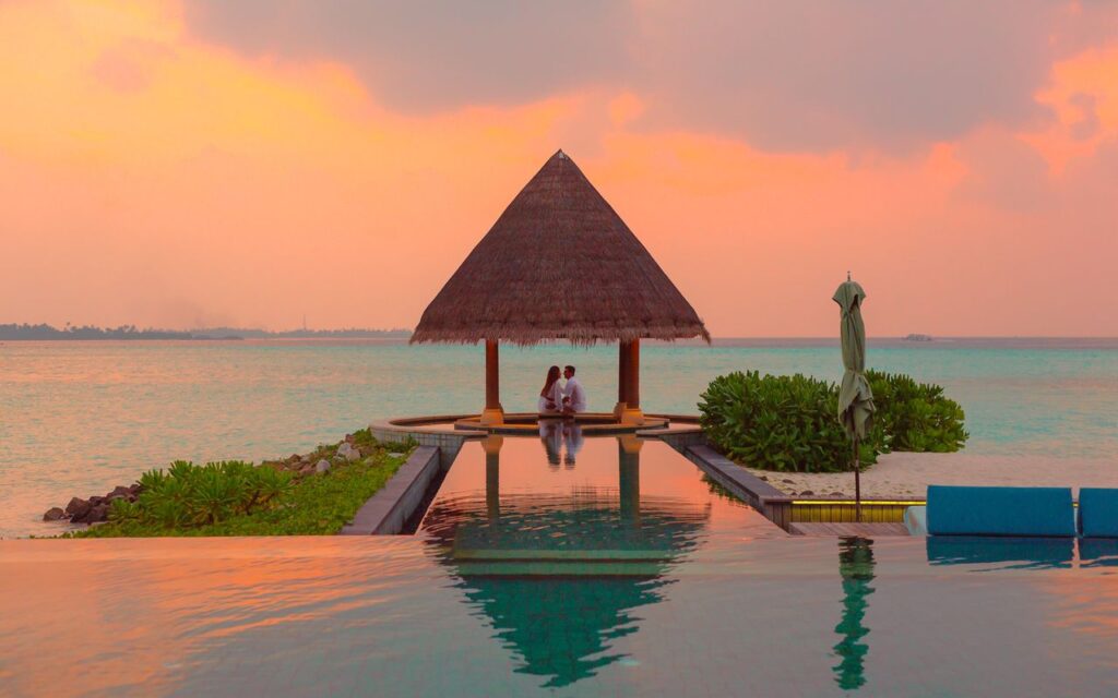 A couple enjoying a romantic moment by the sea and pool under a beach cabana during sunset.